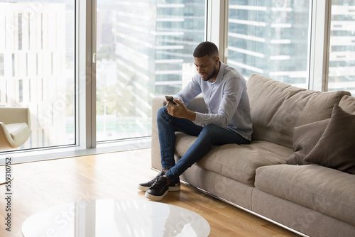 In luxury skyscraper penthouse, young African man use smartphone resting on sofa, spend weekend on internet at home. Modern tech, mobile application usage, electronic services buyer, e-commerce client