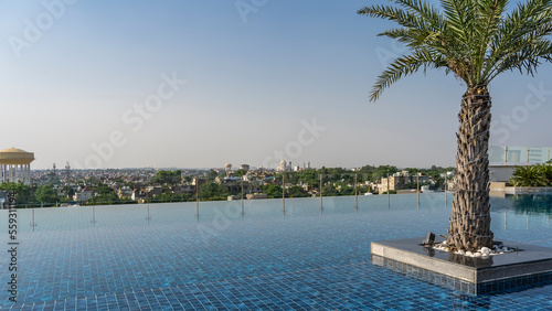 Infinity pool on the roof. The blue tiled bottom is visible through the clear water. Palm tree against the blue sky. The city is visible in the distance. India