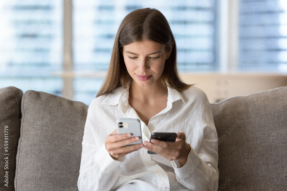 Woman holding two modern smart phones seated on couch, owner of new wireless gadget transfers data, contacts, files and photos from old model of device, compares software, use technology and back up Woman holding two modern smart phones seated on couch, owner of new wireless gadget transfers data, contacts, files and photos from old model of device, compares software, use technology and back up