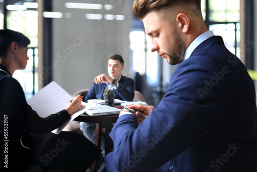 Businessman looks at his wrist watch checking the time. Businessperson sitting a meeting and working at background