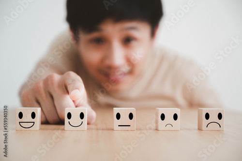 Hand of a asian man and smiley face point to a smiley on wood block cube, Business services rating and satisfaction survey concept.