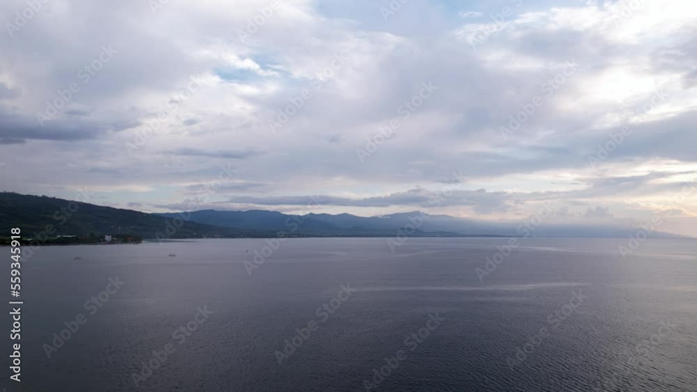 Panoramic view, camera fly at some distance from Lovina Beach, north shore of Bali island, in evening hour. Calm sea water seen below, cloudy sky and dark land far ahead Panoramic view, camera fly at some distance from Lovina Beach, north shore of Bali island, in evening hour. Calm sea water seen below, cloudy sky and dark land far ahead