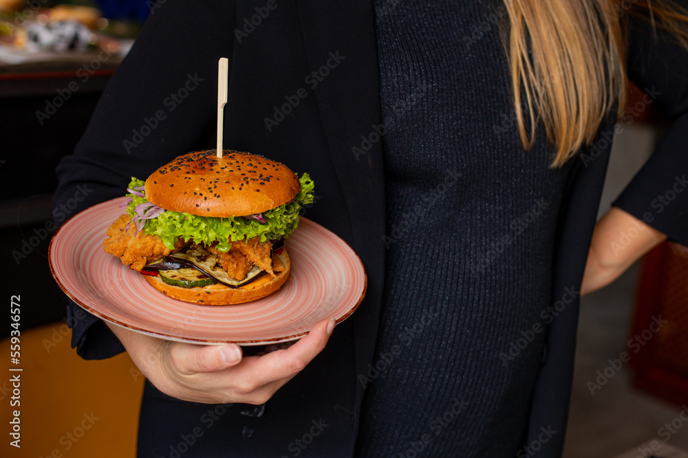 Woman in black clothes hold in hand plate with juicy appetizing chicken burger in public catering. Serving of big caloric sandwich on skewer. Like fast food, self service, restaurant dish, dinner