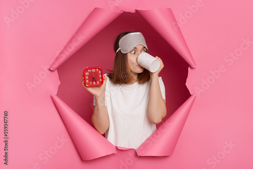 Indoor shot of beautiful brunette young woman wearing white T-shirt and sleeping mask breaking through paper hole of pink background, showing alarm clock and drinking coffee in morning. photo