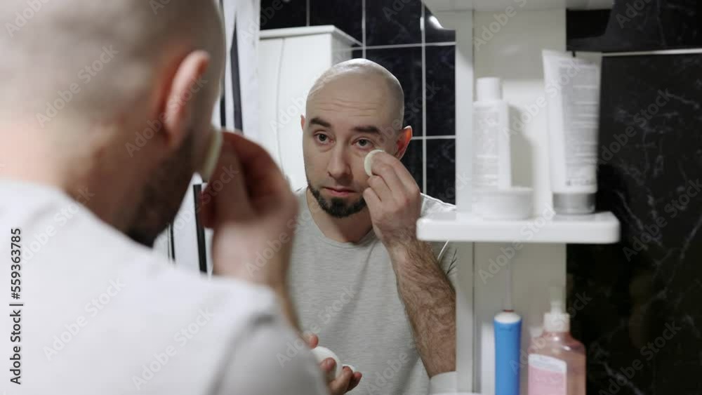 Man's makeup. Portrait of handsome young caucasian bearded bald man using powder at his face and looking at himself in mirror. Man's makeup. Portrait of handsome young caucasian bearded bald man using powder at his face and looking at himself in mirror.