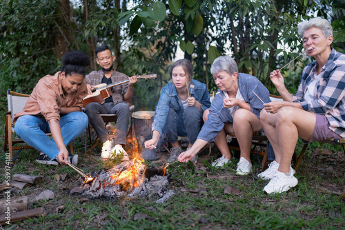 Group of family camper van sitting by fire grilling sausages and singing dancing have fun