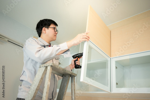 Asian male furniture assembler using electric drill screwdriver on cabinet hinge of glass panel. Interior construction worker man standing on ladder using screwdriving tools for furniture installation
