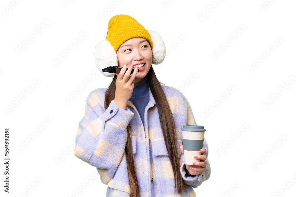 Young Asian woman wearing winter muffs over isolated chroma key background holding coffee to take away and a mobile Young Asian woman wearing winter muffs over isolated chroma key background holding coffee to take away and a mobile