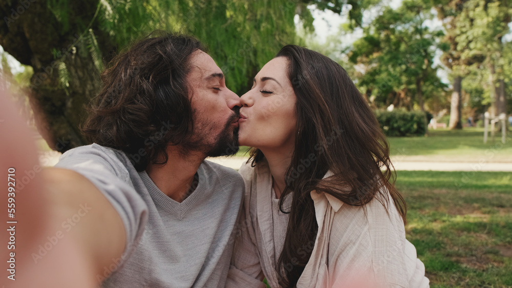 Fototapeta premium Happy couple kissing and taking a selfie sitting on blanket in park