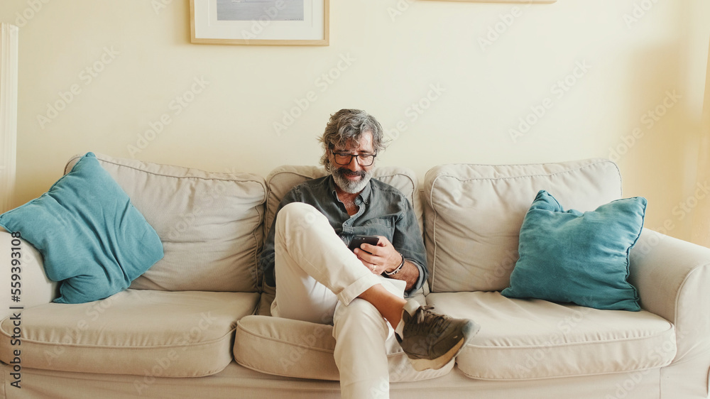 Middle-aged man relaxing on his couch while in home and using his smartphone to text Middle-aged man relaxing on his couch while in home and using his smartphone to text