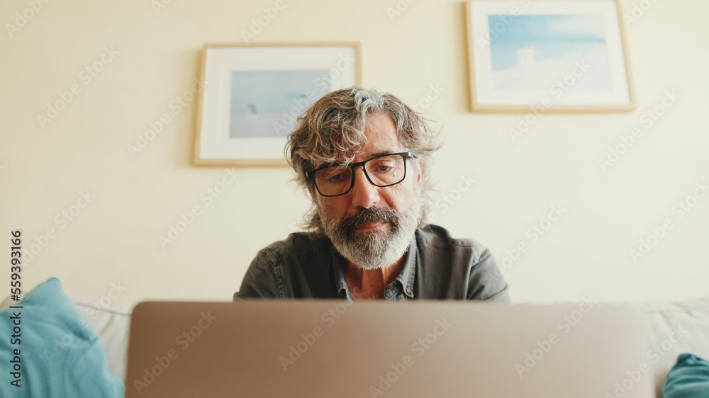 Elderly positive man typing on laptop sitting on sofa in living room Elderly positive man typing on laptop sitting on sofa in living room