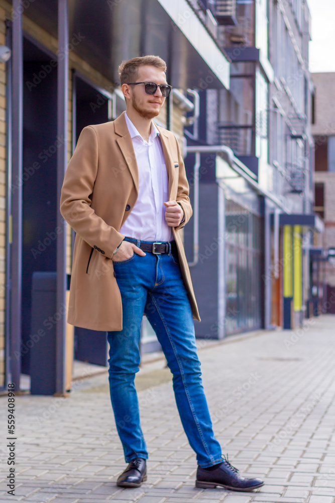 A young man dressed in a beige coat is walking down the street. A young bearded guy with a modern hairstyle and sunglasses on an urban background