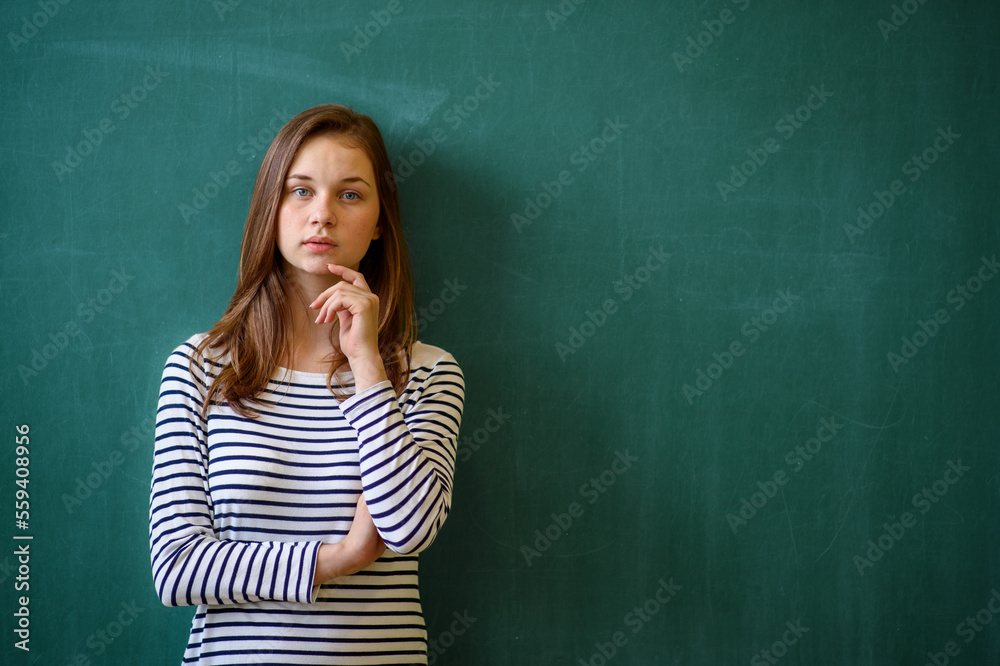Young female high school student standing in front of chalkboard in classroom, looking at camera. Waist up portrait. Young female high school student standing in front of chalkboard in classroom, looking at camera. Waist up portrait.