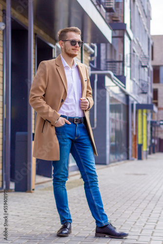 Obraz na płótnie A young man dressed in a beige coat is walking down the street. A young bearded guy with a modern hairstyle and sunglasses on an urban background w salonie