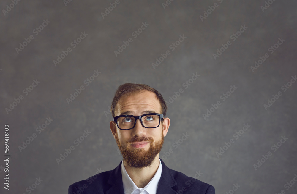 Portrait of unsure male employee with funny dreamy facial expression on gray background. Close up of the face of a man in glasses and suit who looks up dreaming and expecting something.
