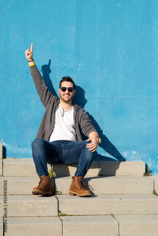 Front view of young bearded man sitting on outdoors staircase, looking away and smiling while raising hand Front view of young bearded man sitting on outdoors staircase, looking away and smiling while raising hand