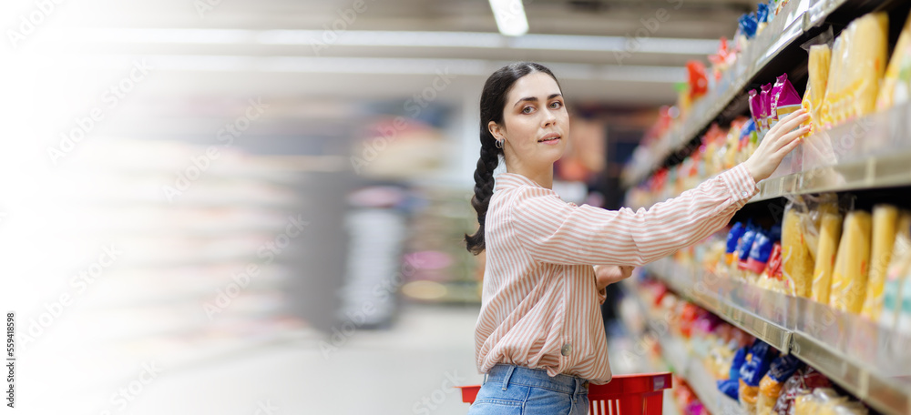Web banner of shopping. Portrait of young smiling beautiful caucasian woman reaches hand to takes food from shelf. Mock up. Copy space. Aisle in background. Concept of consumerism