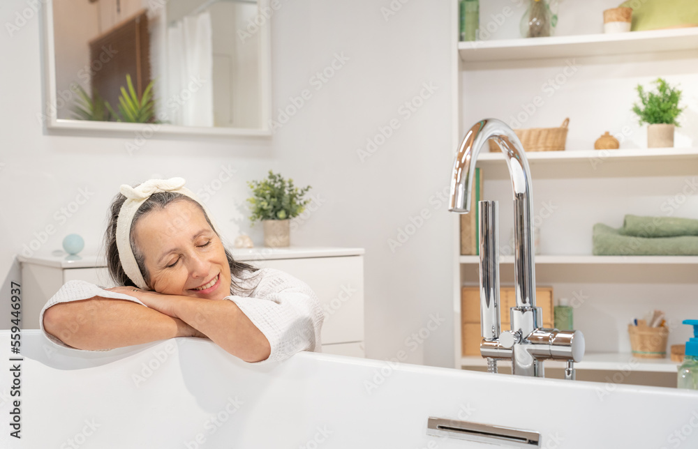 Smiling middle aged woman in bathrobe and headband leaning with crossed arms, relaxing with closed eyes at bathtub. Bodycare spa procedures antiage recreation skin care and beauty cosmetic concept. Smiling middle aged woman in bathrobe and headband leaning with crossed arms, relaxing with closed eyes at bathtub. Bodycare spa procedures antiage recreation skin care and beauty cosmetic concept.