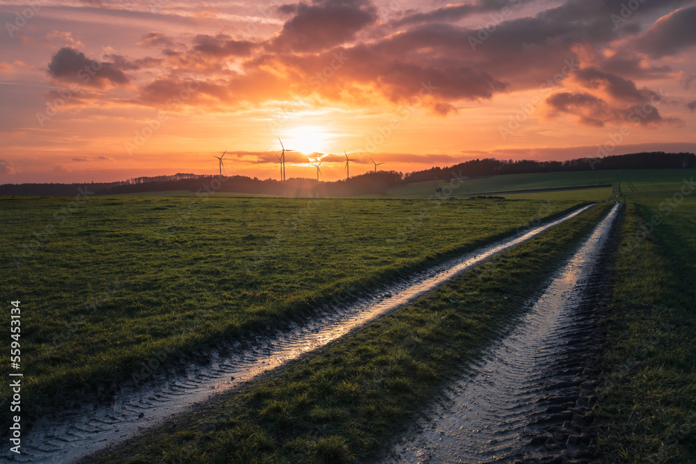 dirt road on the field with the sunset over the wind turbines under the cloudy sky