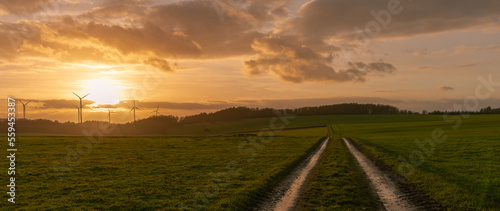 Fototapeta Naklejka Na Ścianę i Meble -  dirt road on the field with the sunset over the wind turbines under the cloudy sky