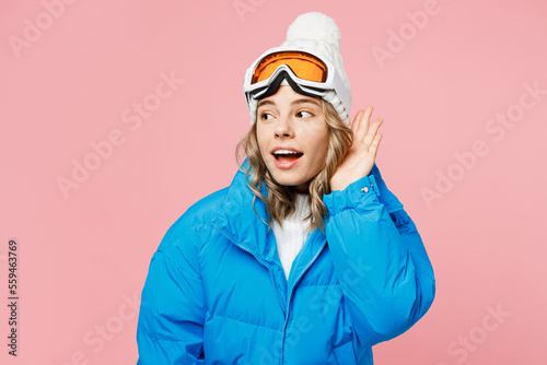 Snowboarder woman in blue suit goggles mask hat ski padded jacket try hear you overhear listen intently isolated on plain pastel pink background Winter extreme sport hobby weekend trip relax concept