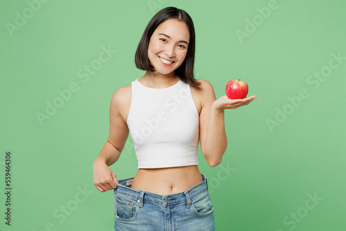 Fotografia Young woman wears white clothes show loose pants on waist after weightloss hold red apple isolated on plain pastel light green background