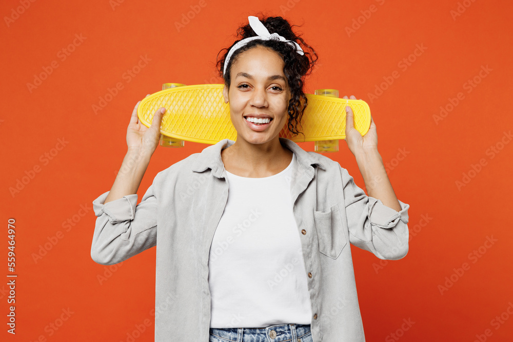 Young smiling fun woman of African American ethnicity she wears grey shirt headband hold behind neck skateboard pennyboard isolated on plain orange background studio portrait People lifestyle concept Young smiling fun woman of African American ethnicity she wears grey shirt headband hold behind neck skateboard pennyboard isolated on plain orange background studio portrait People lifestyle concept