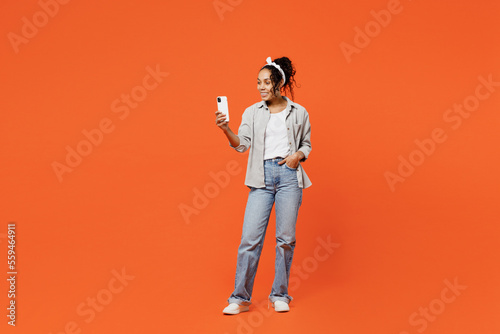 Full body fun young woman of African American ethnicity she wears grey shirt headband hold in hand use mobile cell phone isolated on plain orange background studio portrait. People lifestyle concept.