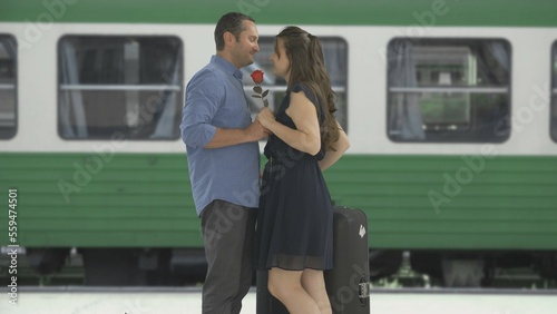 Couple embrace in train station, man waiting woman with red flower