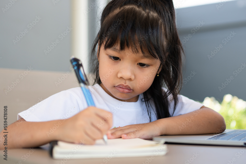 Asian girl student holding a pen for taking notes and doing homework and using the laptop for online learning class at home, Education and distance learning for children Homeschooling concept Asian girl student holding a pen for taking notes and doing homework and using the laptop for online learning class at home, Education and distance learning for children Homeschooling concept