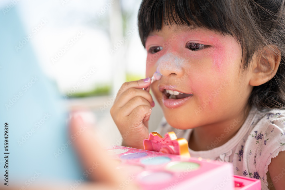 Adorable little child Asian girl paints her mouth with pink children heads and looks in the mirror. A child plays at home in a toy beauty salon. Increase learning development for preschoolers. Adorable little child Asian girl paints her mouth with pink children heads and looks in the mirror. A child plays at home in a toy beauty salon. Increase learning development for preschoolers.