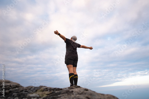 A happy hiker with arm raised up and standing on the rock against blue sky background. 