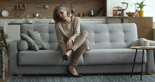 Attractive middle-aged overworked woman goes to sofa sit down taking off her high-heeled shoes to relieve sore, tiredness, discomfort of feet, feels exhausted after hard long workday, rest coming home
