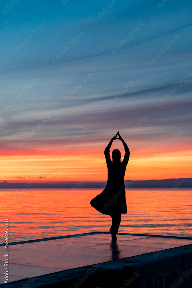 Silhouette of female person doing yoga tree pose by the sea at sunrise or sunset with colourful sky in the background