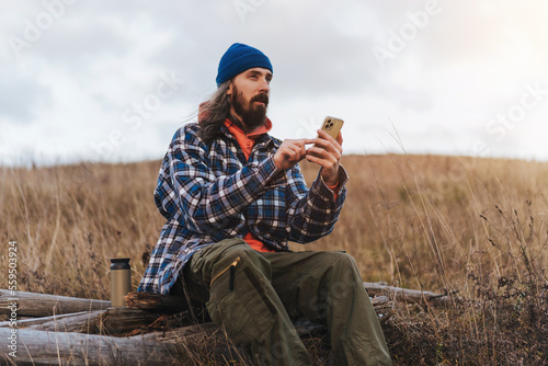 Young bearded male hiker in blue jacket checking the map on smart phone while sitting against blurred green hills during trekking in autumn countryside on sunset.