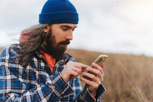 Close up of male hand of bearded male travel photographer in blue jacket holding smart phone while sitting against blurred green hills during trekking in autumn countryside on sunset.