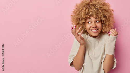 Happy curly haired woman applies medical adhesive plaster for treatment of injuries cuts on skin wears elastic bandage on broken arm dressed in casual beige t shirt isolated over pink background