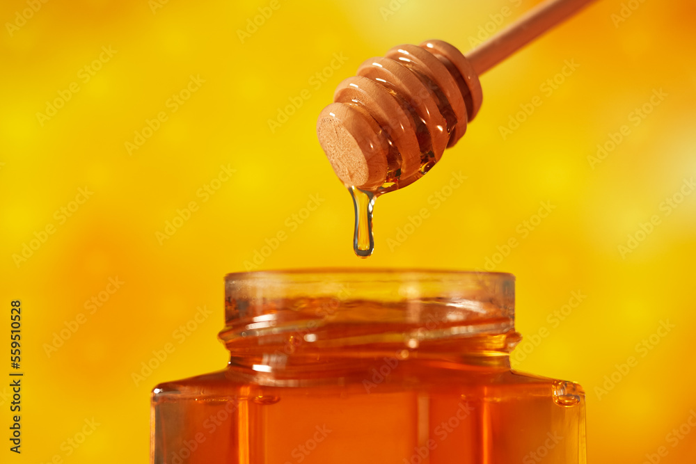 Getting honey from a jar with a wood dipper. Close up view of this healthy food made by bees in nature, acacia tree honey photographed against honeycomb yellow background.