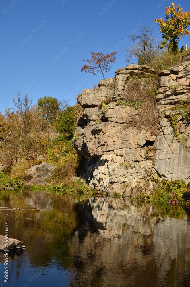 Blue sky above. High rocks. Single trees on rocks. Below is water. Reflection in water.