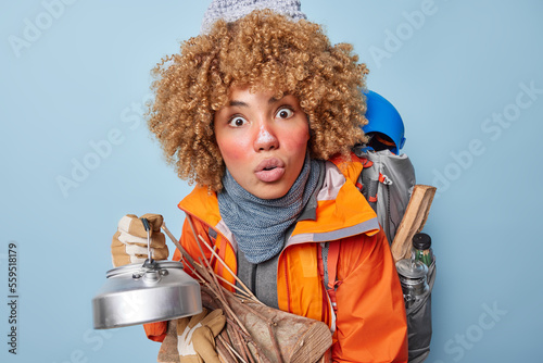 Obraz na płótnie Photo of shocked curly haired woman carries kettle and wood applies protective facial cream on frozen skin poses with rucksack has camping trip isolated over blue background