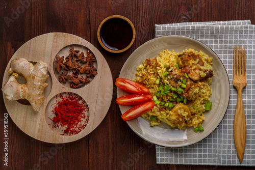 Thai chicken rice garnished with green onions on a wooden table next to a soy sauce napkin and a wooden fork next to anise saffron and ginger.