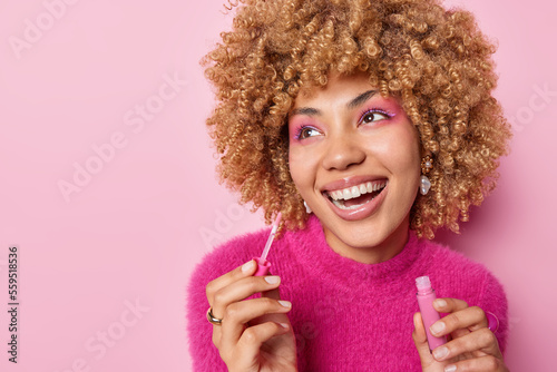 Positive dreamy woman with curly hair applies lip gloss wears vivid makeup smiles broadly has perfect white teeth wears casual pink jumper poses indoor undergoes beauty procedures wants to look nice