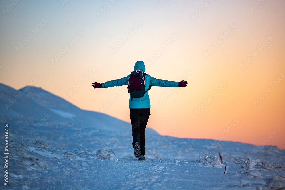 Backpacker girl admires colorful sunset while standing on top of a snowy mountain during cold winter weather Backpacker girl admires colorful sunset while standing on top of a snowy mountain during cold winter weather