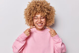 Photo of cheerful curly haired young woman points at perfect white teeth takes care of oral hygiene keeps eyes closed dressed in pink pullover isolated over grey background. Dentistry concept
