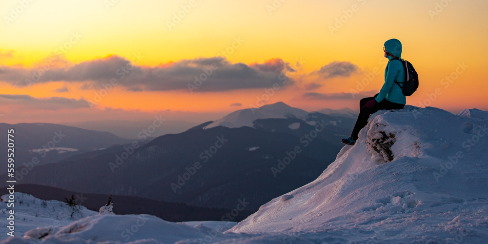 A girl with a backpack sits on an icy mountain top marveling at a colorful sunset during the cold winter weather