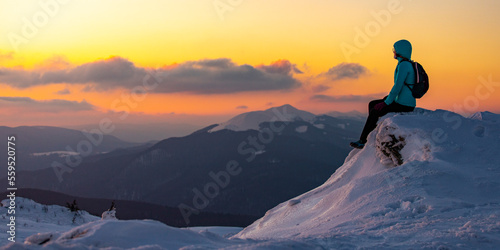 A girl with a backpack sits on an icy mountain top marveling at a colorful sunset during the cold winter weather