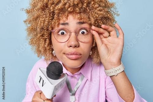 Close up shot of shocked curly woman TV presenter stares impressed through spectacles holds microphone near mouth reports breaking news wears formal pink shirt isolated over blue background. photo