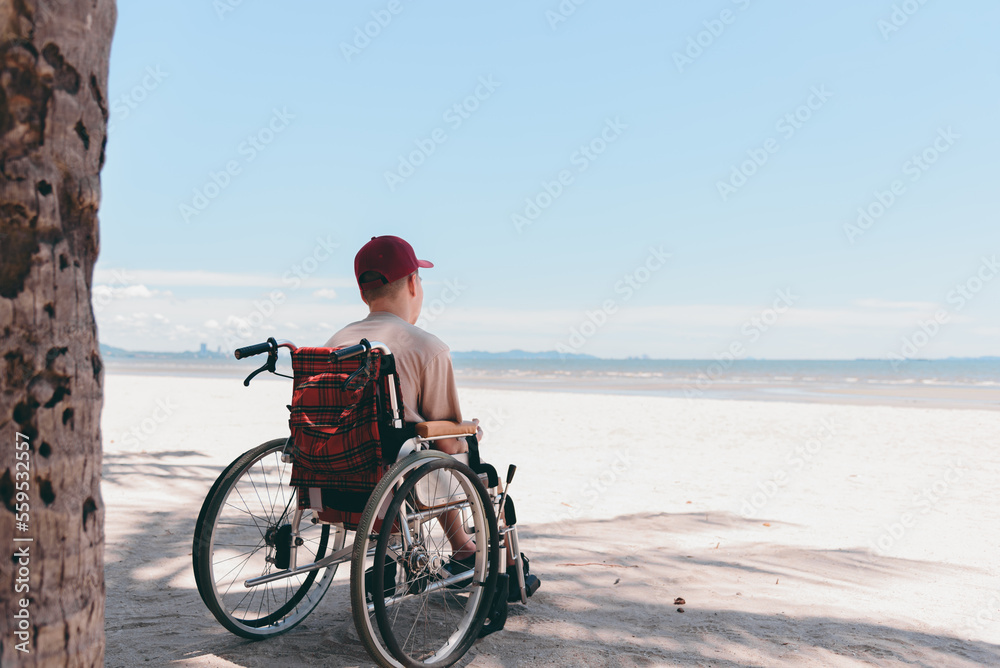 Fototapeta premium Back of young man with disability wearing a red cap on wheelchair, Activity outdoors on the beach background, Vacation on holiday and natural therapy and mental health concept.