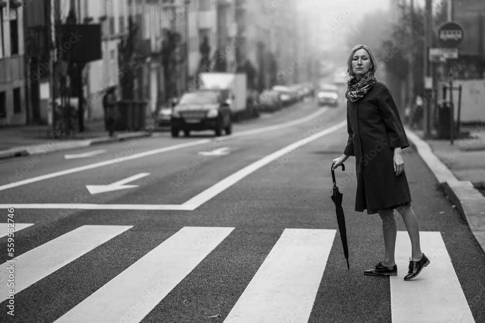 A woman with an umbrella crossing the street in the fog. Black and white photograph. A woman with an umbrella crossing the street in the fog. Black and white photograph.
