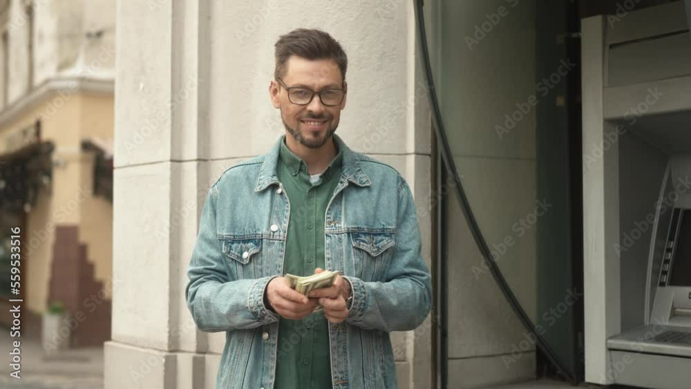 Portrait of cheerful caucasian rich man counting money cashed out from bank credit card using ATM terminal on street. Beautiful happy man looking at camera smiling after financial operations.